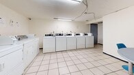 This shared laundry facility is a single-floor space designed for practical, communal use. The main laundry area is the centerpiece of the space, lined with multiple top-loading washing machines along one wall and a row of dryers along the opposite side, providing ample capacity for residents. The machines are all white and uniformly arranged, contributing to a clean, organized appearance. The floors throughout are covered in white ceramic square tiles, lending a bright and easy-to-maintain surface underfoot. Overhead, fluorescent tube lighting runs across the ceiling, ensuring the space is well-lit at all times. The walls are painted white, reinforcing the utilitarian, no-frills aesthetic of the facility. A small alcove area features a round gray table and a blue chair, offering a modest waiting area for users during laundry cycles. Exposed structural elements, including a dark steel column and visible overhead piping, are characteristic of the building's utilitarian construction. The space also includes an exit door with signage, a small trash receptacle, and a utility area with cleaning supplies tucked into a corner. Informational notices are posted on the walls near the machines, providing operational guidance for users. Overall, the facility is straightforward and functional, optimized for efficient shared laundry use.