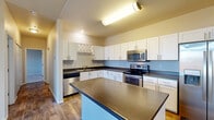 This single-floor residence offers a thoughtfully designed layout that balances functionality and comfort throughout. The kitchen serves as a focal point of the home, showcasing crisp white shaker-style cabinetry, dark laminate countertops, and a full suite of stainless steel appliances including a range, microwave, dishwasher, and French door refrigerator. A central island provides additional prep and dining space, while wood-look plank flooring ties the area together with a warm, clean aesthetic. A decorative wine rack accent adds a charming touch above the sink area. The living area is carpeted and bathed in natural light, with a sliding glass door that opens to a private balcony overlooking a landscaped outdoor setting with mature trees and neighboring greenery. The bedrooms continue the soft carpet flooring and feature generous closet space, including a wide sliding door closet with panel detailing and a spacious walk-in style closet equipped with wire shelving. The bathroom is well-appointed with white cabinetry, dark countertops, wood-look tile flooring, and a full tub and shower combination with white tile surround. Throughout the space, neutral tones, clean lines, and practical finishes create a move-in-ready environment suited for everyday living.