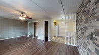 This single-floor unit offers a practical and straightforward layout designed to maximize functionality. The main living area is expansive and open, featuring rich dark hardwood-style flooring and a ceiling fan with integrated lighting. The walls display a distinctive block tile finish, lending a sturdy, textured character to the space. Two windows along the front wall allow natural light to filter in through dark curtain panels, while a wall-mounted HVAC unit provides climate control. An accent wall clad in stacked stone-style material adds a rustic focal point near the entry door.

The living area transitions seamlessly into the sleeping zone, which includes two mirrored sliding-door closets offering generous storage, as well as an additional walk-in closet nearby. A white refrigerator is visible from this area, marking the entrance to the compact kitchen. The kitchen is appointed with warm wood cabinetry, a slate-style tile backsplash, stone-look tile flooring, a coil-burner electric range, over-the-range microwave, and a single-basin sink — all the essentials for everyday cooking. A pendant light above adds warmth to this area.

The bathroom features white square wall tile surrounding a full soaking tub with shower, complemented by painted concrete block walls, and a standard toilet, providing a clean and functional space.