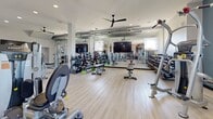 This expansive fitness center offers a generously sized, single-floor exercise space designed to accommodate a wide variety of workout routines. The interior features light wood-style flooring throughout, lofty ceilings fitted with multiple large black ceiling fans, and recessed lighting that ensures the space remains bright and well-ventilated.

The main workout area is thoughtfully organized into distinct training zones. A robust selection of selectorized strength machines and cable systems are arranged across the open floor, providing ample space between equipment for safe and comfortable use. A dedicated free weights section includes a full dumbbell rack and adjustable benches, accompanied by large mirrors along the walls to support proper form. Flat-screen televisions are mounted throughout for entertainment during workouts.

A standout feature is the functional training zone, set against a striking exposed-brick accent wall adorned with fitness-themed wall art. This area includes a multi-functional rig with TRX suspension trainers, battle ropes, medicine balls, and a heavy punching bag, catering to high-intensity and cross-training workouts.

The entry area provides a welcoming transition into the facility, complete with a storage cubby unit, a bench, and dual access doors with exterior views. Teal accent walls add a modern, energetic aesthetic to the overall design, contributing to a motivating and professional fitness environment.