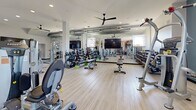 This expansive fitness center offers a generously sized, single-floor exercise space designed to accommodate a wide variety of workout routines. The interior features light wood-style flooring throughout, lofty ceilings fitted with multiple large black ceiling fans, and recessed lighting that ensures the space remains bright and well-ventilated.

The main workout area is thoughtfully organized into distinct training zones. A robust selection of selectorized strength machines and cable systems are arranged across the open floor, providing ample space between equipment for safe and comfortable use. A dedicated free weights section includes a full dumbbell rack and adjustable benches, accompanied by large mirrors along the walls to support proper form. Flat-screen televisions are mounted throughout for entertainment during workouts.

A standout feature is the functional training zone, set against a striking exposed-brick accent wall adorned with fitness-themed wall art. This area includes a multi-functional rig with TRX suspension trainers, battle ropes, medicine balls, and a heavy punching bag, catering to high-intensity and cross-training workouts.

The entry area provides a welcoming transition into the facility, complete with a storage cubby unit, a bench, and dual access doors with exterior views. Teal accent walls add a modern, energetic aesthetic to the overall design, contributing to a motivating and professional fitness environment.