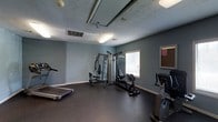 This single-floor fitness space is purpose-built to support a well-rounded exercise regimen. The room is finished with cool-toned blue-gray walls that contribute to a focused and energizing atmosphere. Durable dark rubberized flooring covers the entire space, providing a practical and resilient surface well-suited for physical activity and equipment use.

The room is outfitted with a selection of fitness equipment, including a multi-station cable and weight machine that accommodates a variety of strength training exercises, a treadmill for cardiovascular workouts, and a stationary bike for low-impact cycling. A bulletin board is mounted on one wall, offering a space for notices or posted information.

Natural light filters in through a window, complementing the overhead fluorescent lighting to keep the room well-lit throughout the day. The textured ceiling adds a subtle architectural detail to the otherwise straightforward, functional design. HVAC vents are integrated into the ceiling, ensuring proper climate control within the workout space. Overall, the room is thoughtfully arranged to maximize usable floor area while keeping all equipment accessible and the environment conducive to exercise.