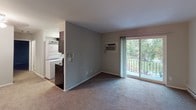 This single-floor unit offers a practical and well-organized layout across several distinct living spaces. The living area is generously sized, finished with wall-to-wall carpeting in neutral tones, and anchored by a sliding glass door that opens to a private balcony overlooking a green outdoor setting. A wall-mounted air conditioning unit provides climate control, complementing the baseboard heating found throughout the carpeted rooms.

The kitchen is designed for efficiency, featuring a galley-style configuration with dark espresso cabinetry, light stone-patterned countertops, and black appliances including an electric range, microwave, and refrigerator. Wood-look flooring runs through the kitchen, and a window allows natural light to brighten the space. In-unit laundry is conveniently integrated into the kitchen area, with a full-size washer and dryer stacked alongside the cabinetry.

The bedrooms are carpeted and spacious, with neutral gray walls, baseboard heating, and windows that frame views of surrounding greenery. Closet space is accessible via bifold doors. The bathroom includes a full tub-shower combination with a curtain rod, a white vanity with a single sink, dark cabinetry below, and tile flooring, with a window providing natural ventilation.