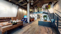 Located at 2200 W Meeker St in Kent, Washington, this two-story commercial space masterfully blends industrial and modern design elements into a cohesive, welcoming environment. The entrance greets visitors with floor-to-ceiling glass walls and doors that flood the foyer with natural light, framing views of the surrounding landscape and lush potted greenery.

The main floor is anchored by wide-plank hardwood flooring, exposed concrete columns, and dramatic dark steel ceiling beams that lend an industrial character throughout. A reception desk with a warm wood finish serves as the central hub, positioned beneath a striking open staircase with black metal railings and wire mesh detailing that leads to the second floor. Distinct seating vignettes populate the main level, including a leather sofa lounge with a wall-mounted fireplace set against a textured reclaimed wood feature wall, as well as a cluster of rich burnt-orange velvet chairs arranged around a television display. Glass-partitioned rooms with bold graphic panels provide semi-private meeting areas. The lower-level lounge area offers a more intimate atmosphere, with exposed ductwork, concrete textures, blue accent chairs, and curated artwork.

Upstairs, an open mezzanine overlooks the main floor, adorned with vibrant artwork and greenery. Throughout the space, carefully selected art pieces, abundant plants, and layered lighting contribute to a refined yet approachable professional ambiance.
