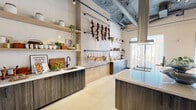 Located at 2141 N Miami Ave in Miami's vibrant Wynwood neighborhood, this single-floor commercial space offers a refined blend of industrial and contemporary design. The open-plan layout is divided into two distinct functional zones seamlessly connected by polished large-format tile flooring throughout.

The expansive kitchen area is a standout feature, showcasing dark wood-grain cabinetry paired with crisp white quartz countertops and a large central island equipped with an integrated sink, induction cooktop, and a professional-grade overhead range hood. Open wooden shelving lines the walls, displaying curated cookware and décor, while an industrial pipe rack suspends an array of copper pots alongside dried botanicals, adding warmth and character. Exposed ductwork and concrete ceilings lend an authentic industrial aesthetic, complemented by a glass-enclosed wine display wall that serves as a dramatic architectural accent.

The dining and event area is equally impressive, bathed in natural light streaming through floor-to-ceiling windows dressed with sheer curtains. A sculptural cluster of gold bowl-shaped pendant lights crowns the space, casting a warm glow over a marble-top dining table surrounded by plush upholstered chairs. A geometric area rug and a curated gallery wall of framed artwork complete this polished, gallery-like atmosphere.