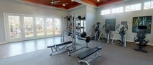 This single-floor fitness center is designed to accommodate a full range of workout needs within a thoughtfully organized layout. The space is divided into distinct functional zones, each tailored to a specific type of exercise. A striking architectural detail unifies the interior: bold orange tray ceilings with recessed lighting and ceiling fans provide both visual interest and practical ventilation throughout the facility.

The cardio zone is outfitted with multiple treadmills, stationary bikes, and elliptical machines, each equipped with individual monitors. High clerestory windows along the walls bring in natural light while maintaining privacy. A multi-functional cable machine and free weight area with a power rack, barbells, and adjustable benches serves as a dedicated strength training section. Wall-mounted mirrors in this area enhance the sense of openness and allow for form monitoring during workouts.

A separate open studio space features wood-look flooring, large wall-to-wall mirrors, and a mounted flat-screen television, making it well suited for yoga, stretching, or group fitness activities. A glass-panel exterior door provides direct outdoor access and additional natural light. The overall aesthetic is clean and modern, with neutral gray walls complementing the bold ceiling accents.