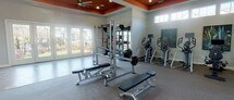 This single-floor fitness center is designed to accommodate a full range of workout needs within a thoughtfully organized layout. The space is divided into distinct functional zones, each tailored to a specific type of exercise. A striking architectural detail unifies the interior: bold orange tray ceilings with recessed lighting and ceiling fans provide both visual interest and practical ventilation throughout the facility.

The cardio zone is outfitted with multiple treadmills, stationary bikes, and elliptical machines, each equipped with individual monitors. High clerestory windows along the walls bring in natural light while maintaining privacy. A multi-functional cable machine and free weight area with a power rack, barbells, and adjustable benches serves as a dedicated strength training section. Wall-mounted mirrors in this area enhance the sense of openness and allow for form monitoring during workouts.

A separate open studio space features wood-look flooring, large wall-to-wall mirrors, and a mounted flat-screen television, making it well suited for yoga, stretching, or group fitness activities. A glass-panel exterior door provides direct outdoor access and additional natural light. The overall aesthetic is clean and modern, with neutral gray walls complementing the bold ceiling accents.