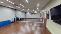 This single-floor fitness studio is thoughtfully designed to accommodate a wide range of exercise disciplines and training styles. The space features rich hardwood flooring that provides both durability and an aesthetically warm foundation underfoot, ideal for dance, barre, yoga, or general fitness routines. A full-length mirrored wall runs along one side of the room, allowing participants to monitor their form and technique during workouts while also creating a sense of openness throughout the space. A wall-mounted ballet barre spans the length of the mirror, offering support for barre exercises, stretching, and balance training. Overhead, a suspended ceiling with recessed linear lighting ensures the room is evenly and brightly illuminated throughout. The studio is equipped with an assortment of fitness accessories, including stability balls in various sizes mounted on the wall, TRX suspension training straps, dumbbells, and a flat-screen display for guided instruction. The walls are finished in a neutral sage and grey palette, contributing to a calm and focused atmosphere. An adjacent corridor and doorway provide easy access to the broader facility, making this studio a functional and inviting space for both individual and group fitness activities.