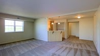 This single-floor residence offers an open-concept layout designed for comfortable, everyday living. The spacious living area is finished with soft wall-to-wall carpeting and benefits from natural light streaming in through a window, creating a bright and welcoming atmosphere. Neutral tones throughout the space lend a clean, understated aesthetic.

At the heart of the floor plan sits a kitchen defined by a curved breakfast bar with light wood laminate detailing, providing a natural gathering point and a seamless visual connection between the kitchen and living spaces. Pendant lighting hangs above the bar, adding a touch of warmth and definition to the area. The kitchen is equipped with standard appliances, including a refrigerator, and is finished with light cabinetry.

Beyond the kitchen, an additional carpeted room extends the living space further, offering flexibility for use as a bedroom, den, or home office. The layout flows intuitively from one area to the next, making the overall footprint feel open and functional. Consistent finishes, including light-colored walls, carpeted flooring in the living areas, and coordinated cabinetry, tie the space together cohesively throughout.