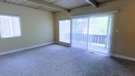This single-floor space showcases a thoughtfully designed interior that blends natural materials with an open, airy atmosphere. The living area is immediately distinguished by its striking exposed wood beam ceiling, which adds warmth and a classic architectural character to the room. Soft, neutral-toned carpeting covers the floors, providing a comfortable and cohesive foundation throughout the space. Large sliding glass doors, dressed with vertical blinds, serve as a prominent feature of the main living area, allowing an abundance of natural light to pour in while offering direct views of the lush greenery outside. A secondary window further enhances the brightness of the room, ensuring the interior feels open and connected to the outdoors. The walls are finished in a soft, warm tone that complements the natural wood elements above. Clean white baseboards provide a crisp contrast and a polished finish to the overall aesthetic. The layout is generously proportioned, offering a versatile and welcoming living environment. The combination of natural textures, ample light, and thoughtful design details creates a calm and inviting ambiance throughout this well-appointed space.