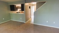 This two-story residence offers a functional and well-connected layout designed for comfortable everyday living. The main level showcases an open-concept design where the living area flows seamlessly into the kitchen, which is visible through a pass-through opening. The kitchen is equipped with dark cabinetry, a range with overhead ventilation, and a light-toned countertop, creating a practical workspace with an efficient use of space. The living area features wall-to-wall carpeting in a warm neutral tone, contributing to a cozy and inviting atmosphere. Soft gray walls carry throughout the main level, lending a calm, cohesive aesthetic to the interior. A staircase leads to the upper floor, positioned alongside a hallway that connects to additional rooms, hinting at the generous layout spread across both levels. Flush-mount ceiling lighting provides consistent illumination throughout. The overall design is straightforward and practical, with clean lines and a neutral palette that allows for versatile interior styling. The two-floor configuration provides a natural separation between living and private spaces, making this a well-organized home suited for a variety of lifestyles.