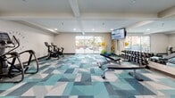 The fitness center at Mayfair Station in El Cerrito, California is a thoughtfully designed, single-floor facility that offers a comprehensive workout environment. The space is anchored by a vibrant geometric-patterned carpet in shades of teal, blue, and gray, lending the room a modern and energetic aesthetic that complements its clean, neutral-toned walls.

The cardio zone features a row of spin bikes, elliptical trainers, and treadmills, all equipped with digital screens for an engaging workout experience. A wall-mounted flat-screen television adds to the entertainment options during exercise sessions. A full-length mirrored wall along one side visually expands the space while allowing users to monitor their form.

The strength training area is well-appointed with a tiered dumbbell rack offering a wide range of weights, a weight bench, medicine balls, and a multi-functional cable machine. Recessed lighting throughout ensures the space is evenly and brightly lit, maintaining a clean and inviting atmosphere.

Large sliding glass doors open directly to an outdoor patio area, allowing natural light to flow in and providing views of the surrounding community. The branded Mayfair Station logo displayed prominently on one wall reinforces the identity of this well-maintained community amenity. A secondary entrance door adds to the accessibility and convenience of the facility.