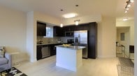 This single-floor residence showcases a thoughtfully designed, open-concept layout that maximizes both functionality and style. The kitchen serves as the heart of the space, featuring bold, dark espresso cabinetry that rises to the ceiling for ample storage, paired elegantly with granite countertops that provide a refined contrast. A central island with a clean white base and matching granite surface offers additional prep space and a natural gathering point. Stainless steel appliances, including a French door refrigerator, built-in microwave, and dishwasher, deliver a polished, contemporary look. Pendant lighting and a flush ceiling fixture illuminate the kitchen warmly, enhancing the inviting ambiance. Light-toned large-format tile flooring flows throughout, lending a sense of openness and continuity to the space. A window above the sink introduces natural light, brightening the kitchen and offering a pleasant connection to the outdoors. The neutral walls and bright, airy palette complement the darker cabinetry, creating a balanced and sophisticated aesthetic throughout this well-appointed single-level residence.