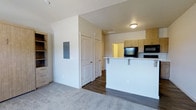 This single-floor space offers a clean, contemporary design with an open-concept layout that connects the living area and kitchen in a seamless, functional flow. The living area is finished with plush carpet flooring, providing a comfortable and inviting atmosphere, while the kitchen and adjoining spaces feature rich dark hardwood-style flooring that adds warmth and contrast to the neutral palette throughout.

The kitchen is outfitted with light wood cabinetry that extends to the upper walls, offering ample storage. A peninsula with a white countertop and brushed hardware serves as both a prep space and a casual dining or entertaining area. Black appliances, including a microwave and additional cooking appliances, are integrated neatly into the cabinetry, contributing to the kitchen's sleek, modern aesthetic.

Crisp white walls and flush-mount ceiling lighting keep the space bright and well-illuminated. A utility panel is mounted on the wall near the entryway corridor, and two interior doors lead to additional rooms, suggesting private quarters accessible from a central hallway. A freestanding shelving unit in a warm wood tone anchors the living area, providing additional storage and display space. Overall, the unit presents a well-organized, modern living environment with practical design details throughout.