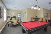 Located at 3100 Hampton Hwy in Yorktown, Virginia, this single-floor community amenity space is designed to serve as a versatile gathering and recreation hub. The open layout seamlessly connects multiple functional zones, creating an inviting atmosphere for leisure and socializing.

At the heart of the space sits a full-size billiards table with a rich red felt surface, illuminated by a stylish pendant light fixture overhead. Surrounding the pool table, multiple seating arrangements invite relaxation, including a pair of tufted leather armchairs, patterned accent chairs, and a glass-top coffee table that anchor a comfortable lounge area.

A dedicated gaming and activity table with cushioned chairs provides space for card games, puzzles, or casual meetings. A built-in media wall with dark wood cabinetry, open shelving, and a large flat-screen television serves as a standout feature, offering additional entertainment options.

Throughout the space, large windows with white horizontal blinds allow generous natural light to filter in, offering views of the surrounding greenery. The warm beige walls, drop-panel ceiling with recessed and pendant lighting, and patterned carpet flooring contribute to a cohesive, comfortable aesthetic. Decorative artwork and plants add subtle character throughout, making this space both functional and welcoming for everyday community use.