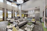 Located at 23400 Kingsland Blvd in Houston (Katy), TX, this single-floor community clubhouse delivers a polished, contemporary environment designed for both socializing and productivity. The space is defined by dramatically soaring ceilings supported by textured stone columns, large windows that flood the interior with natural light, and a warm color palette of grey, taupe, and accent chartreuse green. Patterned modular carpet tiles with vibrant green borders guide movement throughout the open-plan layout. A central lounge area features plush grey sofas, accent chairs, and glass-top coffee tables arranged around a wood-paneled media wall outfitted with multiple flat-screen displays. Adjacent seating vignettes offer additional relaxed gathering spots. The kitchen and bar area is a standout feature, equipped with stainless steel appliances including a full-size refrigerator, a mosaic tile backsplash, open wood shelving, and a generous stone-topped island with modern globe pendant lighting overhead. A dedicated workspace with a desk, computer workstation, and task seating is tucked near the large windows. Oversized drum pendant fixtures and globe pendant lights provide layered illumination throughout, contributing to the space's refined, hospitality-inspired ambiance.