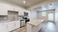 This efficiently designed single-floor, one-bedroom, one-bathroom unit offers a thoughtful layout with modern finishes throughout. The kitchen serves as a central highlight, featuring crisp white shaker cabinets, granite countertops, a subway tile backsplash, a central island with additional counter space, and a full suite of stainless steel appliances including a refrigerator, range, microwave, and dishwasher. Wood-look luxury vinyl plank flooring runs throughout the main living areas, adding warmth and durability.

The open living area is bright and welcoming, with natural light filtering in through windows fitted with blinds and direct access through a glass-paneled door. Neutral tones on the walls complement the flooring and create a clean, versatile aesthetic.

The primary bedroom is generously proportioned, offering plush carpeting, a ceiling fan, and a window that brings in natural light. It connects to a dedicated walk-in closet equipped with wire shelving and hanging rods for ample storage. The bathroom is finished with a white vanity topped with granite counters, wood-look flooring, and a tiled tub and shower surround, maintaining the unit's consistent modern aesthetic throughout.