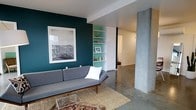 This modern single-floor space showcases a compelling blend of industrial and contemporary design. The living area immediately draws the eye with its striking deep teal accent wall, which serves as a bold backdrop for a large-format black-and-white framed artwork. Polished concrete floors run throughout, lending an industrial character while reflecting light and creating a sense of openness. A signature exposed concrete column anchors the space, adding a raw, structural aesthetic that complements the clean white walls and ceiling.

The living area is anchored by a mid-century modern gray sofa set atop a layered combination of a woven jute rug and a patterned area rug, adding warmth and texture to the cool concrete surfaces. A sleek glass-top coffee table completes the seating arrangement.

Built-in shelving, finished in a complementary teal tone, is seamlessly integrated into the wall adjacent to the accent feature, providing both functional storage and a display area for décor. The open layout connects the living space to additional rooms beyond, offering a fluid sense of flow throughout the interior. The overall ambiance is modern, curated, and thoughtfully designed, balancing industrial elements with warm, inviting touches.