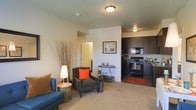 This single-floor residence offers a thoughtfully designed open-concept layout that seamlessly connects the living area and kitchen, creating a cohesive and functional living environment. The living space features plush carpeted flooring and is furnished with comfortable seating, making it an inviting area for relaxation. Warm, neutral-toned walls are complemented by flush-mount ceiling lighting that provides consistent illumination throughout.

The kitchen is a standout feature, showcasing rich dark espresso cabinetry that extends to the upper walls, offering generous storage space. Black appliances, including a range, microwave, and dishwasher, lend a sleek, uniform aesthetic to the kitchen. A subtle transition between the carpeted living area and the kitchen adds visual definition to the space.

A partial wall subtly separates the living and kitchen zones while maintaining the open feel of the floor plan. A doorway visible from the living area hints at additional private spaces beyond, such as a bedroom or bathroom. The overall ambiance is modern yet welcoming, with a neutral color palette that provides a versatile backdrop for a variety of interior styles. The layout makes efficient use of space, offering a comfortable and practical single-floor living experience.