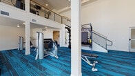 This two-story fitness center is designed to accommodate a wide range of workout needs in a modern, well-equipped environment. The ground floor serves as the primary exercise area, boasting soaring double-height ceilings that create an open, energizing atmosphere. Large windows allow natural light to pour in, illuminating a generous layout filled with strength training stations, cable machines, free weight racks with a full dumbbell selection, TRX suspension systems, and adjustable benches. A staircase with a sleek metal railing connects to the upper level, which hosts a row of ellipticals, stationary bikes, and additional cardio equipment, along with views down to the main floor below. Treadmills are positioned near windows on the lower level, offering exterior views during workouts. A dedicated children's play room provides a bright, welcoming nook furnished with a small table and chairs, colorful toys, and framed artwork on the walls — ideal for caregivers working out nearby. An additional open, carpeted multipurpose room offers flexible space for group classes or stretching. Throughout both floors, a striking dark and teal geometric patterned carpet ties the space together, complementing the clean white walls and modern recessed lighting.