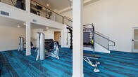 This two-story fitness center is designed to accommodate a wide range of workout needs in a modern, well-equipped environment. The ground floor serves as the primary exercise area, boasting soaring double-height ceilings that create an open, energizing atmosphere. Large windows allow natural light to pour in, illuminating a generous layout filled with strength training stations, cable machines, free weight racks with a full dumbbell selection, TRX suspension systems, and adjustable benches. A staircase with a sleek metal railing connects to the upper level, which hosts a row of ellipticals, stationary bikes, and additional cardio equipment, along with views down to the main floor below. Treadmills are positioned near windows on the lower level, offering exterior views during workouts. A dedicated children's play room provides a bright, welcoming nook furnished with a small table and chairs, colorful toys, and framed artwork on the walls — ideal for caregivers working out nearby. An additional open, carpeted multipurpose room offers flexible space for group classes or stretching. Throughout both floors, a striking dark and teal geometric patterned carpet ties the space together, complementing the clean white walls and modern recessed lighting.