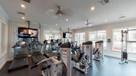 This single-level fitness center in Houston, TX (77049) offers a thoroughly equipped workout environment designed to support a wide range of exercise routines. The space is bright and open, with large windows lining multiple walls that invite generous natural light and provide views of the surrounding outdoor area. Ceiling fans are positioned throughout, contributing to comfortable airflow during workouts.

The cardio zone is well-stocked with True treadmills, elliptical machines, and stationary bikes, several of which are arranged to face wall-mounted flat-screen televisions for entertainment during exercise. A large mirror wall further enhances the sense of space while allowing users to monitor their form.

The strength-training area features a variety of Body-Solid resistance machines targeting different muscle groups, a cable cross machine, adjustable benches, and a full rack of dumbbells. Durable rubber flooring covers the workout areas, providing both safety and comfort underfoot.

The interior design is polished and tasteful, with white crown molding, recessed lighting, and framed abstract artwork on the walls, lending the space a refined aesthetic. An adjacent room, visible through interior windows and a doorway, suggests additional community amenities nearby. Overall, the fitness center is well-maintained, functional, and thoughtfully designed.