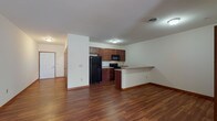 This single-floor unit offers a well-organized open-concept layout designed for comfortable everyday living. Rich, warm-toned hardwood-style flooring extends throughout the main living areas, creating a cohesive and inviting atmosphere. The spacious living area provides generous room for a variety of furniture arrangements and benefits from natural light filtering in through the windows.

The kitchen is positioned adjacent to the living space and features rich medium-toned cabinetry with ample upper and lower storage. Light-colored countertops provide a functional workspace, and black appliances—including a refrigerator, electric range, and over-the-range microwave—are included. A peninsula counter with an extended overhang defines the boundary between the kitchen and living area, offering additional prep space or casual seating.

A dedicated utility area accommodates a stacked washer and dryer unit, adding notable convenience. The bedroom is carpeted for comfort, with a window that allows natural light and outdoor views of surrounding greenery, contributing to a calm, restful environment. Throughout the unit, white walls, consistent trim work, and neutral tones create a clean, versatile backdrop suitable for a range of interior styles.