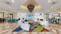 This single-story clubhouse is a comprehensively amenitized community space designed to blend sophisticated style with functional leisure. The centerpiece of the facility is a expansive lobby lounge distinguished by soaring coffered ceilings and a dramatic multi-tiered chandelier composed of cylindrical glass and metal elements in jewel tones. The floors throughout feature a striking herringbone-patterned wood design, and the seating area is anchored by a bold, multicolored area rug in teal, magenta, and chartreuse beneath a curved sectional sofa flanked by elegant white accent chairs. Textured stone wall cladding and vibrant artwork contribute to the space's lively yet refined aesthetic, while abundant natural light pours in through large black-framed windows and glass doors.

Adjacent areas include a well-equipped fitness center outfitted with treadmills, ellipticals, and strength equipment. A game room, labeled "Hilarity Hall," features a billiard table and additional gaming options. A lounge area with a tile-surround fireplace and mounted display provides a cozy retreat. The kitchen area showcases bold plum and white cabinetry, granite countertops, and commercial-grade refrigerators stocked with beverages. A café nook with a coffee station, live-edge dining table, and pendant lighting completes the social amenity offerings, with direct access to an outdoor patio.