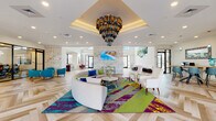 This single-story clubhouse is a comprehensively amenitized community space designed to blend sophisticated style with functional leisure. The centerpiece of the facility is a expansive lobby lounge distinguished by soaring coffered ceilings and a dramatic multi-tiered chandelier composed of cylindrical glass and metal elements in jewel tones. The floors throughout feature a striking herringbone-patterned wood design, and the seating area is anchored by a bold, multicolored area rug in teal, magenta, and chartreuse beneath a curved sectional sofa flanked by elegant white accent chairs. Textured stone wall cladding and vibrant artwork contribute to the space's lively yet refined aesthetic, while abundant natural light pours in through large black-framed windows and glass doors.

Adjacent areas include a well-equipped fitness center outfitted with treadmills, ellipticals, and strength equipment. A game room, labeled "Hilarity Hall," features a billiard table and additional gaming options. A lounge area with a tile-surround fireplace and mounted display provides a cozy retreat. The kitchen area showcases bold plum and white cabinetry, granite countertops, and commercial-grade refrigerators stocked with beverages. A café nook with a coffee station, live-edge dining table, and pendant lighting completes the social amenity offerings, with direct access to an outdoor patio.