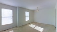 This three-floor property offers a clean, neutral aesthetic defined by light-toned walls and soft carpeted flooring. The image showcases a well-proportioned room dressed in crisp white walls that enhance the sense of openness and airiness. Natural light filters through a double-hung window fitted with horizontal blinds, casting warm sunlight across the carpeted floor and contributing to the room's bright, welcoming atmosphere. A classic chandelier with candelabra-style arms serves as an elegant focal point overhead, adding a traditional touch to the otherwise understated space. The smooth white ceiling and simple white baseboard trim maintain a cohesive, clean finish throughout. The room's generous proportions and uncluttered layout provide a versatile canvas suitable for a variety of uses. With three floors in total, the property offers ample living space distributed across multiple levels, accommodating a range of functional needs. The overall ambiance is light, calm, and move-in ready, with timeless design elements that appeal to a broad range of occupants.