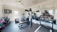 Located in Jackson, TN, this dedicated fitness room is a compact yet fully functional private gym designed to support a comprehensive workout routine. The space features a vaulted ceiling with crown molding and recessed lighting, lending an open, airy feel that belies its modest footprint. Ceiling fans provide continuous air circulation throughout workouts.

The room is outfitted with durable rubber flooring ideal for heavy equipment use and high-impact activity. Floor-to-ceiling mirrors line one wall, visually expanding the space while allowing users to monitor form during exercise. Natural light streams in through multiple windows, offering views of the surrounding neighborhood and keeping the atmosphere bright and inviting.

The fitness area is equipped with a full cable machine/multi-station strength trainer, a treadmill, an elliptical, a weight bench, a medicine ball, and a full rack of dumbbells in varying weights. A wall-mounted flat-screen television is positioned above the dumbbell rack for entertainment during workouts. A water cooler station near the door ensures convenient hydration. A fire extinguisher is also on-site for safety.

The clean, white walls and refined architectural details—including crown molding and a paneled ceiling accent—give this gym a polished, upscale character that sets it apart from a standard workout room.