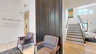 Located at 920 Belmont Ave in North Haledon, NJ, this contemporary amenity center offers a well-appointed, single-floor layout designed for community living and recreation. The welcoming entrance lobby features soaring vaulted ceilings, large format tile flooring, and curated artwork — including a striking mural referencing "The Origin of Molly" — setting a modern, gallery-like tone from the moment you arrive. The spacious lounge area is bathed in natural light through floor-to-ceiling windows, with light hardwood-style flooring, plush seating arrangements, and an accent wall clad in richly textured wood slat paneling. A recreation room stands out with its dramatic angled ceiling, full wood-paneled feature wall, and a vibrant billiards table, offering an energetic social hub. The well-equipped fitness center provides a comprehensive range of cardio and strength training equipment, with dynamic wall murals, ample natural light, and durable rubberized flooring throughout. A fully outfitted kitchenette with dark wood cabinetry, mosaic tile backsplash, and generous counter space supports gatherings and events. A secondary strength training room with a multi-station cable machine completes the fitness offerings. The space seamlessly blends sophisticated design with practical amenities.