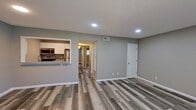 This single-floor space presents a clean, modern aesthetic defined by consistent grey-toned walls, crisp white trim, and durable wood-look laminate flooring that runs throughout. The open living area is bright and welcoming, featuring recessed lighting and a large window dressed with vertical blinds that allows natural light to fill the room. A pass-through window connects the living space directly to the kitchen, which is equipped with stainless steel appliances — including a refrigerator, microwave, and range — set against white cabinetry and granite-style countertops, creating a functional and stylish cooking area. A hallway leads from the main living space to the additional rooms, maintaining a logical and efficient layout. The bedroom is a comfortably sized, private retreat with its own recessed light fixture and a window with blinds that invites natural light. The bathroom is straightforward and well-maintained, featuring a full tub-shower combination in white, a toilet, a vanity, and grey textured walls that complement the overall neutral palette. Throughout the space, textured ceilings, visible HVAC vents, and consistent flooring tie each room together into a cohesive, move-in-ready unit.