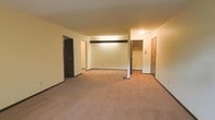 This single-floor space is characterized by an open layout with wall-to-wall carpeting in a warm neutral tone, providing a cohesive and understated foundation throughout. The walls are finished in a clean cream color, contributing to a bright and simple aesthetic. A structural beam accent runs horizontally across the upper portion of the main living area, adding a subtle architectural detail to the otherwise straightforward design.

Multiple doorways are visible from the main living area, indicating access to additional rooms and functional spaces. A mirrored closet with visible shelving and hanging rod is accessible from the main area, offering practical storage solutions. A second closet or utility door is positioned nearby, further enhancing storage options. An additional doorway on the opposite side leads to what appears to be a hallway connecting further rooms.

The space is illuminated by ceiling-mounted light fixtures, providing consistent indoor lighting throughout. Dark baseboards contrast with the lighter walls, framing each room with a defined border. Overall, the space presents a functional, no-frills layout suited for practical everyday living.