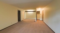 This single-floor space is characterized by an open layout with wall-to-wall carpeting in a warm neutral tone, providing a cohesive and understated foundation throughout. The walls are finished in a clean cream color, contributing to a bright and simple aesthetic. A structural beam accent runs horizontally across the upper portion of the main living area, adding a subtle architectural detail to the otherwise straightforward design.

Multiple doorways are visible from the main living area, indicating access to additional rooms and functional spaces. A mirrored closet with visible shelving and hanging rod is accessible from the main area, offering practical storage solutions. A second closet or utility door is positioned nearby, further enhancing storage options. An additional doorway on the opposite side leads to what appears to be a hallway connecting further rooms.

The space is illuminated by ceiling-mounted light fixtures, providing consistent indoor lighting throughout. Dark baseboards contrast with the lighter walls, framing each room with a defined border. Overall, the space presents a functional, no-frills layout suited for practical everyday living.