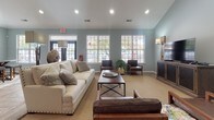 Located at 5080 Sandy Dr in High Point, North Carolina, this single-floor community space is thoughtfully designed to provide a comfortable and stylish gathering environment. The interior is unified by soft blue-gray walls, wood-look tile flooring in a herringbone pattern, and consistent decorative accents that create a warm, cohesive ambiance throughout.

The main lounge area is spacious and inviting, featuring a plush white sofa, leather seating, and a sturdy coffee table centered beneath recessed lighting. Large multi-pane windows line the walls, flooding the room with natural light and offering views of the surrounding outdoor area. The open layout flows seamlessly into a dedicated dining space, where a substantial bar-height table with upholstered stools rests atop a boldly patterned area rug. A decorative mirror with flanking sconce lamps adds an elegant focal point.

Adjacent to the dining area, a wet bar station features crisp white cabinetry, a smooth countertop, and a striking mosaic tile backsplash in neutral earth tones. The entryway area offers a comfortable leather accent chair and a gallery-style wall display of ornate circular wooden medallions, adding artistic character to the arrival experience. Throughout, the décor blends rustic and contemporary elements for a refined yet approachable community space.