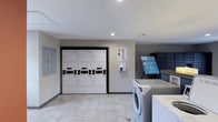 This well-appointed shared laundry facility offers a clean, functional environment designed to meet the everyday needs of its users. The space features a bank of stacked, high-capacity washer and dryer units arranged neatly along one wall, providing ample laundry capacity. The machines are equipped with digital payment terminals, allowing for easy and cashless operation. Light-toned tile flooring runs throughout, contributing to a bright and hygienic atmosphere, while recessed lighting in the white ceiling ensures the room is well-lit at all times. Along the opposite side, the room includes front-loading washing machines, offering additional laundry options. A notable feature is the set of secure package lockers, ideal for convenient parcel storage and retrieval. Informational signage is displayed on the walls, keeping users informed about machine operation and upcoming amenities. The overall design is practical and modern, with neutral gray walls and a streamlined layout that maximizes the usable space. This facility reflects a commitment to providing residents with a convenient, up-to-date laundry experience in a tidy, well-maintained setting.