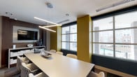 This single-floor professional meeting space is thoughtfully designed with a contemporary aesthetic that balances functionality and visual appeal. The centerpiece of the main conference room is a generously sized, light-toned rectangular table surrounded by upholstered chairs featuring a distinctive geometric pattern, providing comfortable seating for a large group. Overhead, a striking angular linear pendant light fixture serves as a focal point, casting clean, even illumination across the workspace.

The room's color palette combines deep navy blue and warm olive-gold accent walls against a neutral white ceiling, creating a bold yet cohesive atmosphere. Large windows along one wall flood the space with natural light while offering views of the surrounding urban environment. A wall-mounted flat-screen television is positioned above a credenza along one wall, accommodating presentations and video conferencing needs, with a conference phone conveniently placed on the table.

Glass partition walls with slim metal framing divide the space into distinct zones, maintaining an open and transparent feel while defining separate meeting areas. Abstract artwork adorns one wall, adding a creative element to the professional setting. Patterned carpet tiles and recessed lighting throughout contribute to a polished, well-appointed environment suited for collaborative work.