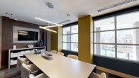This single-floor professional meeting space is thoughtfully designed with a contemporary aesthetic that balances functionality and visual appeal. The centerpiece of the main conference room is a generously sized, light-toned rectangular table surrounded by upholstered chairs featuring a distinctive geometric pattern, providing comfortable seating for a large group. Overhead, a striking angular linear pendant light fixture serves as a focal point, casting clean, even illumination across the workspace.

The room's color palette combines deep navy blue and warm olive-gold accent walls against a neutral white ceiling, creating a bold yet cohesive atmosphere. Large windows along one wall flood the space with natural light while offering views of the surrounding urban environment. A wall-mounted flat-screen television is positioned above a credenza along one wall, accommodating presentations and video conferencing needs, with a conference phone conveniently placed on the table.

Glass partition walls with slim metal framing divide the space into distinct zones, maintaining an open and transparent feel while defining separate meeting areas. Abstract artwork adorns one wall, adding a creative element to the professional setting. Patterned carpet tiles and recessed lighting throughout contribute to a polished, well-appointed environment suited for collaborative work.