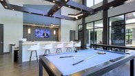 This contemporary amenity lounge is designed to impress, combining entertainment and social functionality within a single, open-concept space. The room's most striking architectural feature is its soaring ceiling, accented by bold dark cross-beam detailing and a combination of recessed lighting and geometric pendant fixtures that illuminate the space with a sleek, modern glow.

At the forefront of the room sits a professional-grade pool table with a light blue felt surface, serving as a natural centerpiece. Adjacent to it, a foosball or game table adds an additional entertainment option. Along one wall, a stylish bar-height counter lined with modern white stools faces a wall-mounted flat-screen television, creating an ideal gathering spot for social events or casual lounging.

The space benefits from floor-to-ceiling glass walls and large black-framed sliding doors that connect seamlessly to an outdoor area, flooding the interior with natural light and providing an open, airy atmosphere. The overall aesthetic is clean and contemporary, combining dark structural accents, light-toned surfaces, and hardwood-style flooring for a polished, upscale feel.