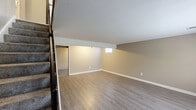 Located at 1210 NW Kent Ct in Blue Springs, MO, this two-story home offers a thoughtfully laid-out floor plan across two levels. The main floor serves as the heart of the home, featuring a combined dining and kitchen area with white cabinetry, a speckled countertop, black appliances, and light wood-style flooring. Striking exposed dark wood beams accent the soaring vaulted ceiling, adding architectural character to the open living space, which is bathed in natural light from large windows. The full bathroom on this level features a white vanity with a quartz-style countertop, modern brushed nickel fixtures, and wood-style flooring. Both bedrooms are carpeted and well-sized, with the primary bedroom benefiting from vaulted ceilings and natural light from multiple windows. The second bedroom includes a window with a neighborhood view. The lower level provides additional versatile living space with wood-style flooring, neutral painted walls, and a small window for light, along with a utility room and pantry. An attached garage with concrete flooring and built-in storage panels completes the home. Multiple closets and a hallway add to the practical storage throughout.
