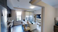 This single-floor residence showcases a thoughtfully designed open-concept layout that seamlessly integrates the living and dining areas into one cohesive, functional space. The interior is finished with warm hardwood-style flooring that runs throughout, complemented by neutral-toned walls that create a clean, modern aesthetic.

The living area is anchored by a comfortable sofa and accented with a glass coffee table, while a wall-mounted flat-screen television sits above a dedicated media zone. A lean ladder-style shelving unit provides stylish open storage alongside the media wall, adding both function and character to the space.

Adjacent to the living area, a compact dining table with upholstered chairs offers an intimate setting for meals or casual gatherings. Multiple windows dressed with roller shades and curtain panels allow abundant natural light to pour in, creating a bright and airy ambiance throughout. The windows also provide views of the surrounding urban streetscape and a balcony beyond, further extending the sense of openness.

Recessed lighting and flush-mount ceiling fixtures ensure the space is well-lit at all times of day. A built-in track system on the ceiling adds a modern architectural detail. The overall design balances comfort and contemporary style, making it a welcoming and highly livable space.