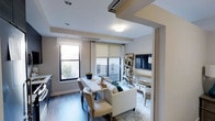 This single-floor residence showcases a thoughtfully designed open-concept layout that seamlessly integrates the living and dining areas into one cohesive, functional space. The interior is finished with warm hardwood-style flooring that runs throughout, complemented by neutral-toned walls that create a clean, modern aesthetic.

The living area is anchored by a comfortable sofa and accented with a glass coffee table, while a wall-mounted flat-screen television sits above a dedicated media zone. A lean ladder-style shelving unit provides stylish open storage alongside the media wall, adding both function and character to the space.

Adjacent to the living area, a compact dining table with upholstered chairs offers an intimate setting for meals or casual gatherings. Multiple windows dressed with roller shades and curtain panels allow abundant natural light to pour in, creating a bright and airy ambiance throughout. The windows also provide views of the surrounding urban streetscape and a balcony beyond, further extending the sense of openness.

Recessed lighting and flush-mount ceiling fixtures ensure the space is well-lit at all times of day. A built-in track system on the ceiling adds a modern architectural detail. The overall design balances comfort and contemporary style, making it a welcoming and highly livable space.