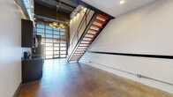Located at 727 N Fourth St in Columbus, Ohio, this two-story live/work space offers a compelling blend of industrial design and modern finishes. The main level makes an immediate impression with its soaring ceilings, polished concrete floors, and exposed structural elements including steel beams and corrugated metal ceiling panels. A full-glass garage door floods the entry area with natural light, while a bold brass chandelier adds a refined contrast to the raw aesthetic. The centerpiece of the main floor is a dramatic open-tread staircase with warm wood steps framed by a black steel perforated metal railing — a true architectural statement. The kitchen is visible from the main living area and is outfitted with stainless steel appliances. Painted concrete block walls throughout the lower level reinforce the industrial character of the space. Upstairs, the upper level offers a more intimate setting with hardwood flooring, housing the primary bedroom and access to both bathrooms. The two full bathrooms are identically appointed with floor-to-ceiling large-format gray tile, glass-enclosed tub/shower combinations, dark wood vanities with granite countertops, and sleek modern fixtures. A closet and hallway complete the upper level. This space delivers a distinctive urban aesthetic suited for creative or flexible living arrangements.
