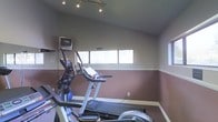 This single-floor fitness room is a purpose-built exercise space designed to support a healthy, active lifestyle. The room features a two-tone wall treatment, with a warm reddish-brown lower half and a neutral upper half, giving the space a defined and energetic atmosphere. Track lighting is mounted on the ceiling, providing bright, focused illumination throughout the workout area. A row of horizontal windows along one wall allows natural light to filter in, creating an open and airy feel despite the room's compact footprint. The space is equipped with multiple cardio machines, including a treadmill, an elliptical trainer, and what appears to be an additional stationary cardio unit, offering a variety of workout options. A wall-mounted television is positioned at an easy-to-view angle, allowing users to stay entertained during their sessions. The flooring is covered in a practical, low-pile carpet suited for fitness use. Electrical outlets are conveniently placed along the walls for powering devices and equipment. Overall, this fitness room delivers a clean, functional, and motivating environment well-suited for daily cardio workouts.