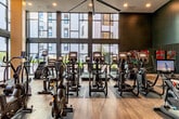 Located at 1350 S Bascom Avenue in San Jose, CA, this two-story fitness center delivers a well-appointed exercise experience across a generously sized layout. The ground floor serves as the primary exercise area and features soaring double-height ceilings, expansive floor-to-ceiling windows that flood the space with natural light, and wide-plank hardwood flooring. This level is equipped with an extensive array of cardio machines, including treadmills, ellipticals, and stationary bikes, all arranged to maximize both functionality and views to the outdoors. Large ceiling fans overhead enhance air circulation throughout. A sleek open staircase with metal railings connects the two floors, creating a visually open and modern transition between levels. The upper floor houses a second dedicated exercise room featuring a more intimate layout with strength training machines and free weights, including a full rack of kettlebells and dumbbells, surrounded by floor-to-ceiling mirrors and windows. Additionally, a flexible studio room on the ground level offers a carpeted, open floor plan with glass partition walls, mirrored surfaces, and a modern ceiling fan, ideal for yoga, stretching, or group classes. The facility's contemporary design is unified by a dark teal and charcoal color palette, recessed lighting, and a clean, professional ambiance throughout.