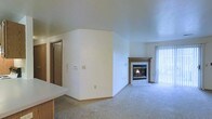 This single-floor residence offers a straightforward and functional layout designed for comfortable everyday living. The main living area is spacious and open, finished with neutral wall-to-wall carpeting that lends a soft, understated feel throughout. A standout feature of the living space is the fireplace, framed by a classic white tile surround and a warm wood mantle, serving as a natural focal point for the room. Natural light flows generously into the space through a large sliding glass door dressed with vertical blinds, offering both brightness and privacy. The sliding door provides direct access to an outdoor area, enhancing the sense of openness. The clean white walls throughout create a bright, neutral canvas well-suited for a variety of décor styles. Interior doors with warm wood tones add a grounding contrast to the light walls and carpeting. The layout includes defined transition spaces, with doorways leading to additional rooms, suggesting a practical and organized floor plan. Overall, the space presents a clean, move-in-ready environment with classic residential finishes and an inviting, no-frills ambiance ideal for a variety of occupants.