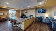 This single-floor communal space is thoughtfully designed to serve multiple functions, combining a full kitchen, casual dining, and a tech-equipped work area within one open layout. The kitchen is outfitted with full-size black appliances, including a refrigerator and range topped by a stainless steel range hood, complemented by gray cabinetry, a light-toned countertop, and a peninsula with bar seating. A double sink and compact appliances such as a coffee maker and microwave add everyday convenience.

Adjacent to the kitchen, a dedicated workstation area features desktop computers and wall-mounted monitors, providing a functional space for computing and business tasks. The dining area offers a mix of round and oval tables paired with orange-cushioned chairs, creating a welcoming environment for casual meals or social gatherings. Decorative sheer fabric with string lights adds a warm, festive touch along the large windows.

Throughout the space, dark hardwood-style flooring contrasts with warm tan walls and a drop tile ceiling with recessed lighting. Expansive windows and glass doors line one side of the room, offering views of and access to an outdoor patio furnished with seating and a fireplace.