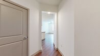 This thoughtfully designed single-floor residence combines modern aesthetics with functional living. Rich, dark hardwood-style flooring flows throughout the main areas, connecting a welcoming entry hallway to the open living and kitchen spaces. The hallway leads past interior doors into a bright, airy living area fitted with a ceiling fan and natural light from slatted blinds.

The kitchen is a standout feature, boasting white shaker-style upper cabinetry, sage-toned lower cabinetry, light quartz countertops, a tile backsplash, stainless steel appliances including a range and over-the-range microwave, and open shelving for added storage and display. A peninsula-style counter extends the workspace and serves as a natural divider between the kitchen and living areas.

The bedroom offers plush carpeting, dual windows that bring in abundant natural light, and a ceiling fan for comfort. The bathroom is elegantly appointed with a wide quartz vanity, white cabinetry, a large framed mirror, dark subway tile in the walk-in glass shower, and hardwood-style flooring that maintains cohesion throughout. The overall palette of crisp whites, warm wood tones, and soft grays creates a clean, contemporary ambiance throughout the space.