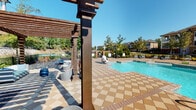 This outdoor amenity area is thoughtfully designed to offer a full resort-style experience within a residential community setting. At its centerpiece sits a generously sized swimming pool with crystal-clear blue water, accessible via stainless steel entry rails and surrounded by an expansive checkerboard-patterned paver deck in warm earth tones. Pool-adjacent lounge chairs line the perimeter, providing ample space for sunbathing under open skies.

Adjacent to the pool, a striking wooden pergola structure serves as the social heart of the outdoor space. Its dark-stained timber beams cast dramatic shadow patterns across the paver hardscape below, creating a comfortable, partially shaded retreat. Beneath the pergola, modern rounded lounge chairs and accent side tables in cool blues and greys are arranged to encourage casual gathering and conversation.

Lush landscaping borders the space throughout, featuring ornamental grasses, decorative shrubs, and mulched garden beds that add texture and color. A stacked stone retaining wall and wrought iron fencing define the boundaries of the space while maintaining an open, airy feel. The multi-building community backdrop, mature trees, and abundant natural light contribute to an inviting, well-maintained outdoor environment suited for year-round enjoyment.